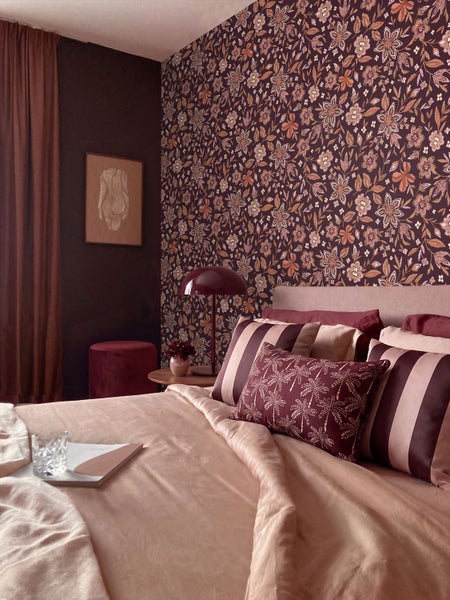 Gemütliches Schlafzimmer mit floraler Mustertapete, Bett mit dekorativen Kissen, Tischleuchte und schweren Vorhängen.