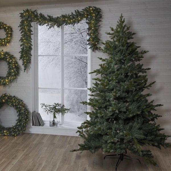 Weihnachtliche Szene mit Tannenbaum, beleuchteter Girlande und Kränzen vor einem Fenster mit Winterlandschaft.