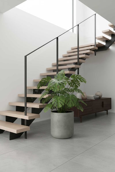 Monstera-Pflanze in grauem Pflanztopf vor einer modernen Holztreppe mit schwarzem Geländer in einem hellen, minimalistischen Wohnraum.