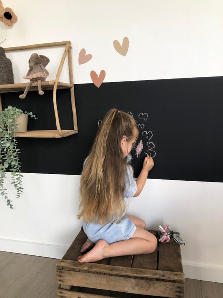 Mädchen zeichnet mit Kreide auf eine Wandtafel in einem Kinderzimmer. Dekorative Elemente wie Herzen und ein Regal mit Spielzeug sind sichtbar.