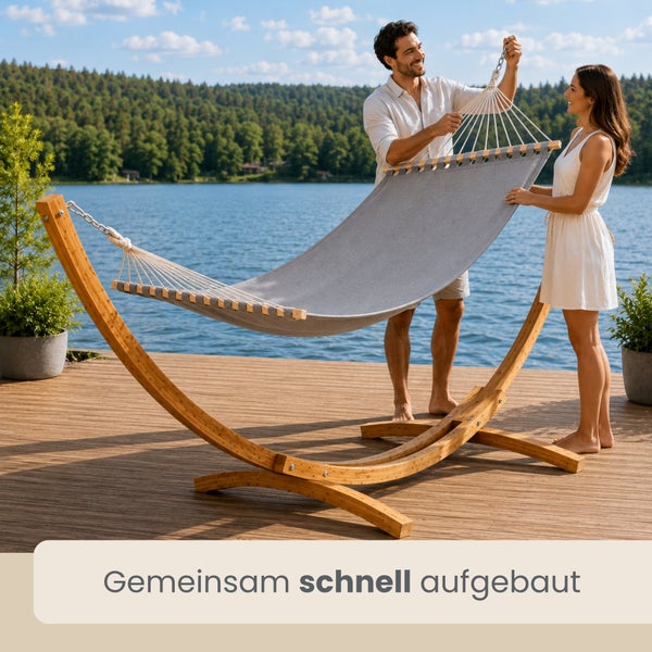 Ein Mann und eine Frau montieren eine graue Hängematte mit einem gebogenen Gestell aus Holz auf einer Terrasse an einem See.