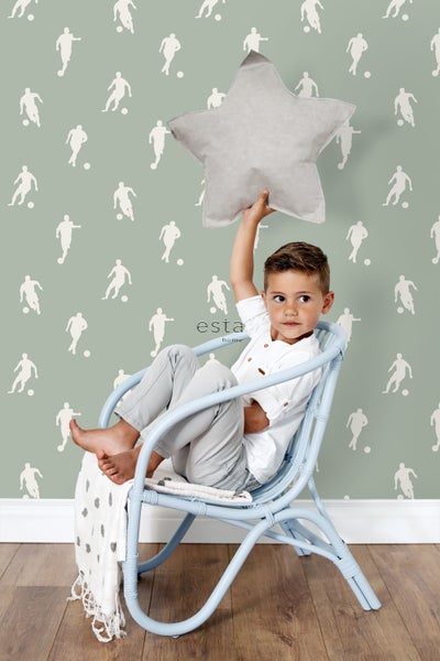 Ein Junge sitzt in einem Sessel vor einer Tapete mit Fußballmotiven und hält ein sternförmiges Kissen.