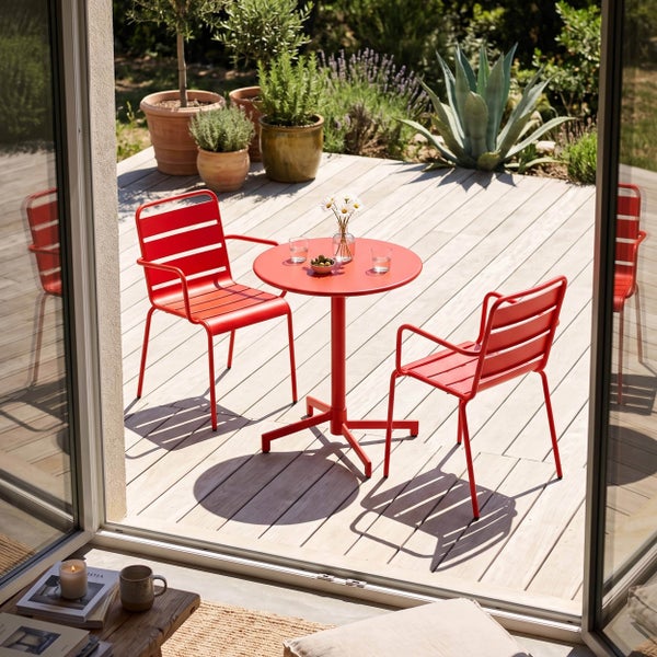 Rotes Metall-Bistrosset mit einem runden Tisch und zwei Stühlen mit Armlehnen auf einer Holzterrasse mit verschiedenen Topfpflanzen.