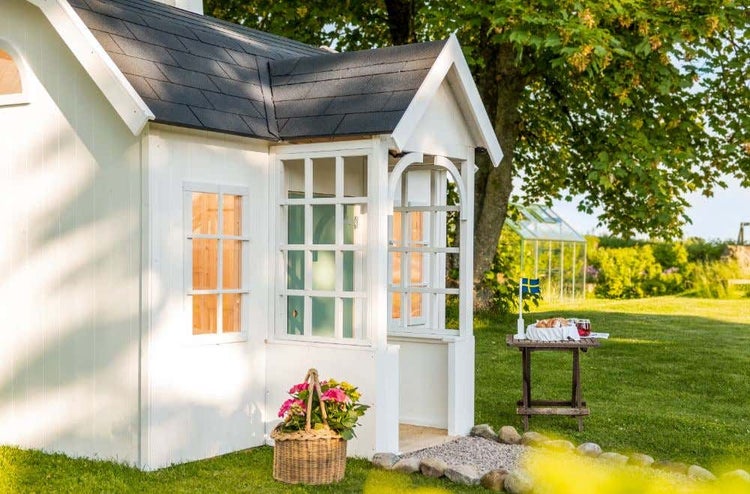 Weißes Gartenhaus aus Holz mit dunklen Schindeln und Sprossenfenstern in einem hellen Garten mit Rasen und Bäumen.