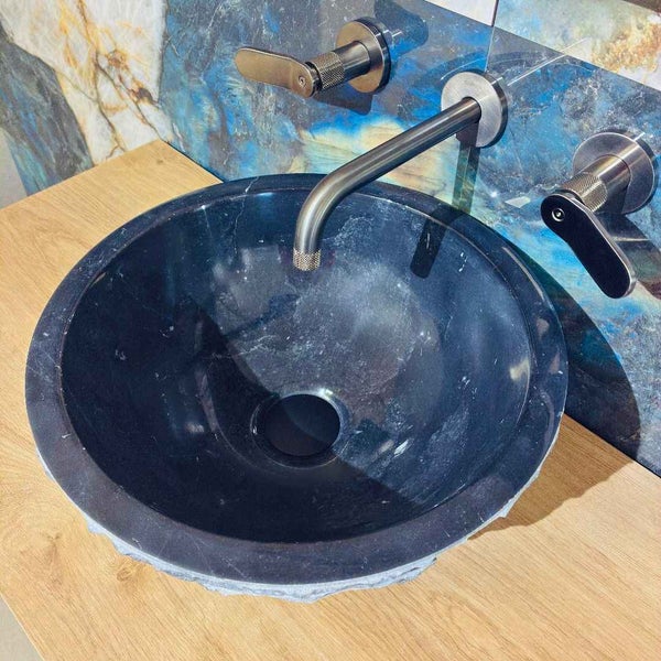 Rundes Aufsatzbecken aus schwarzem Stein auf Holzplatte mit Wandarmatur vor blau gemaserten Fliesen.