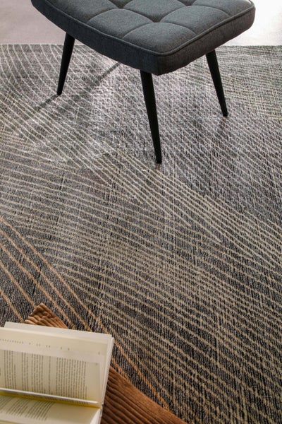 Strukturierter flachgewebter Teppich mit grafischem Linienmuster unter einem Polsterhocker und einem aufgeschlagenen Buch.