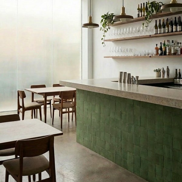 Moderne Bar mit grüner Fliesentheke und Holztischen in einem hellen, offen gestalteten Innenraum mit Hängeleuchten und offenen Holzregalen.