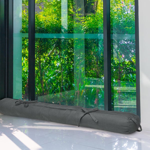 Grauer Zugluftstopper aus Stoff vor einem bodentiefen Fenster auf weißem Fliesenboden.
