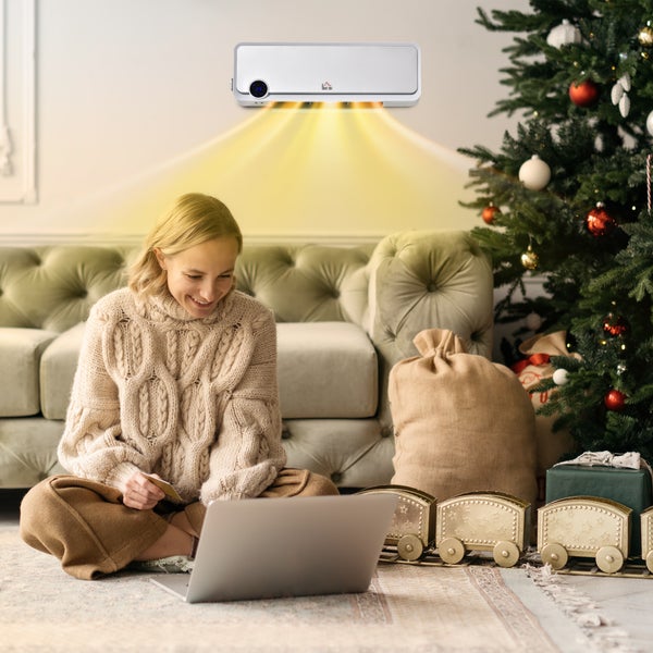 Wandmontiertes Elektroheizgerät strahlt Wärme in ein helles, weihnachtlich dekoriertes Wohnzimmer mit Sofa, Weihnachtsbaum und Frau am Laptop ab.