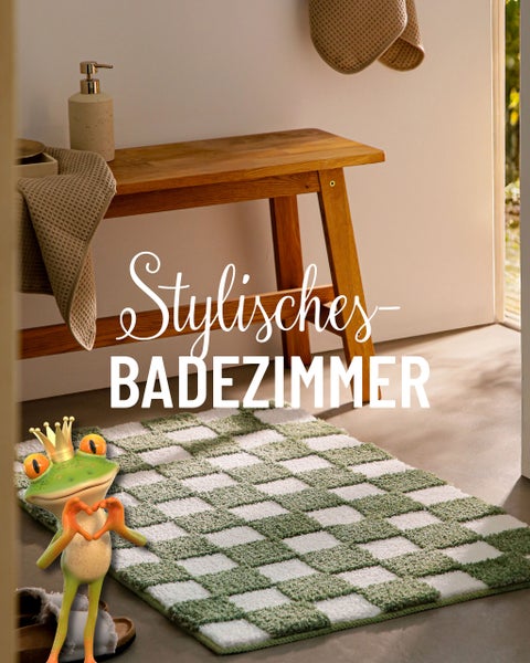 Helles Badezimmer mit Holzbank, Handtüchern und karierter Badematte. Ein gekrönter Frosch steht im Vordergrund.