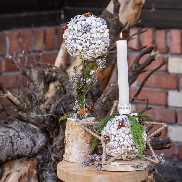 Weihnachtliche Tischdekoration mit einer Kugel aus Zapfen und Eicheln auf einem Holzstamm und einem Kerzenhalter mit Stern aus Jutekordel.