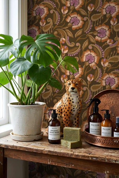 Wohnraumgestaltung mit Monstera-Pflanze in weißem Topf, Leopardenfigur und Pflegeprodukten vor einer floralen Paisley-Tapete.