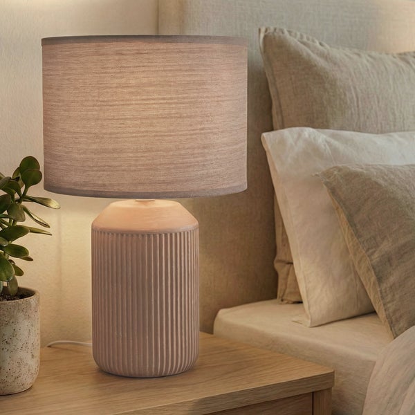 Moderne Tischleuchte mit geriffeltem Keramikfuß und textilem Lampenschirm auf einem Holznachtisch neben einem Bett.