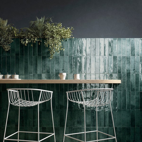 Wand mit glänzenden grünen rechteckigen Fliesen und einer Bar aus Holz mit zwei weißen Drahtstühlen und Zimmerpflanzen im modernen Interieur.