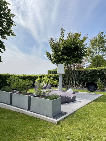 Moderne Gartenlounge mit grauen rechteckigen Pflanzkübeln, einem Sitzsack, einer weißen Außenleuchte und einer schwarzen Feuerschale auf einer Terrasse.