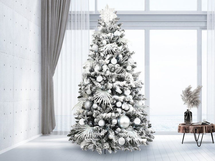 Ein künstlicher, verschneiter Weihnachtsbaum, geschmückt mit Kugeln und dekorativen Elementen, steht in einem hellen, modernen Wohnraum.