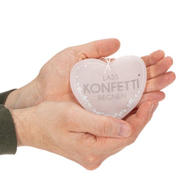 Zwei Hände halten einen lichtdurchlässigen Deko-Herzanhänger mit der Aufschrift Lass Konfetti regnen und weißen Konfetti-Details.