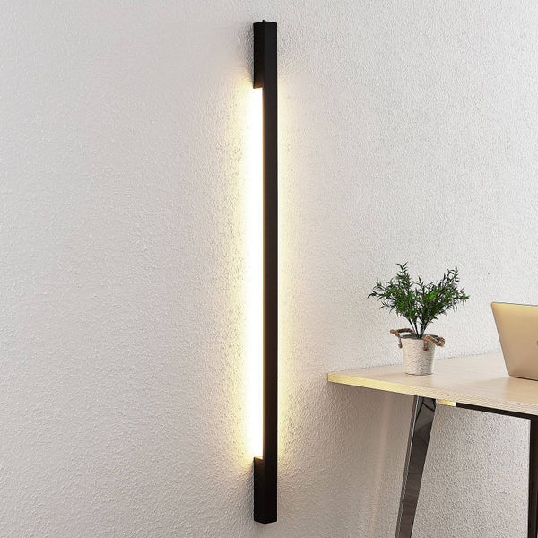Moderne schwarze LED-Wandleuchte in Stabform mit indirektem Licht neben einem Schreibtisch im minimalistischen Stil.