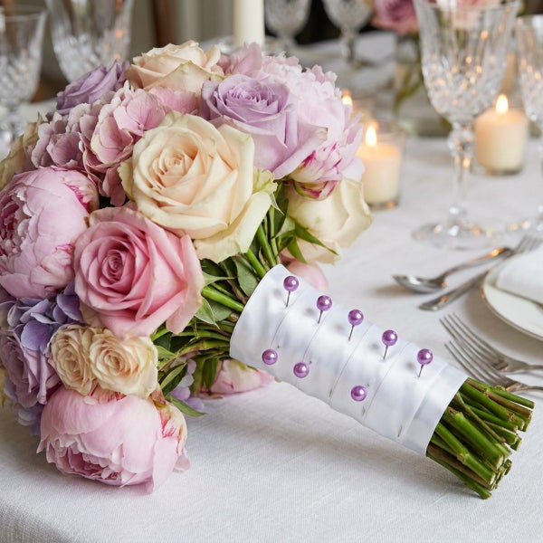 Ein Blumenstrauß aus Rosen und Pfingstrosen, gebunden mit einem weißen Band und Stecknadeln mit Perlenkopf auf einem festlich gedeckten Tisch.