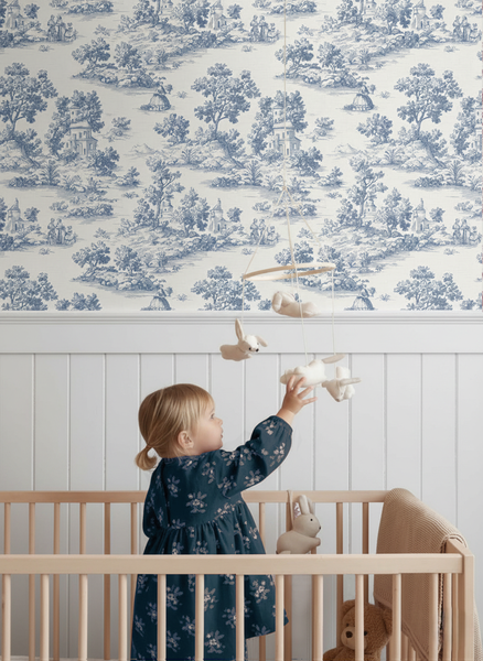 Tapete mit blauem Toile-de-Jouy-Muster in einem Kinderzimmer über weißer Wandtäfelung mit einem Kind im Holzbett.