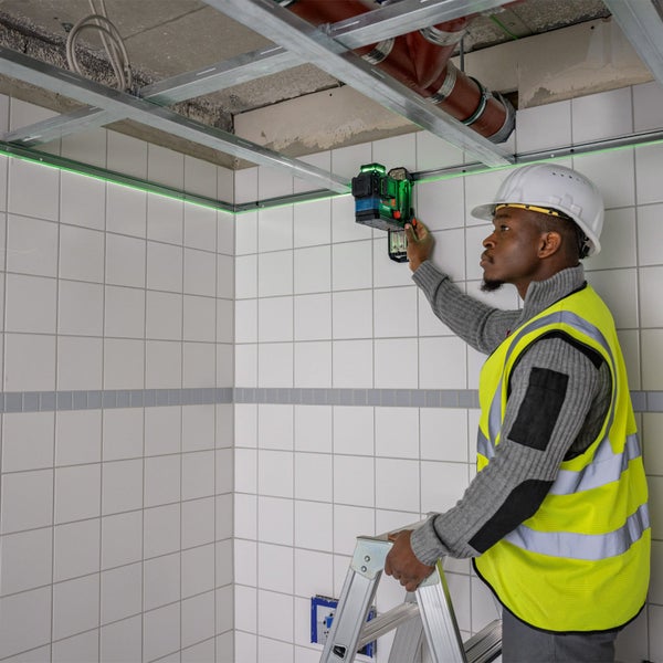 Bauarbeiter mit Schutzhelm und Warnweste auf einer Leiter nutzt einen Kreuzlinienlaser zur Ausrichtung von Metallprofilen an einer Fliesenwand.
