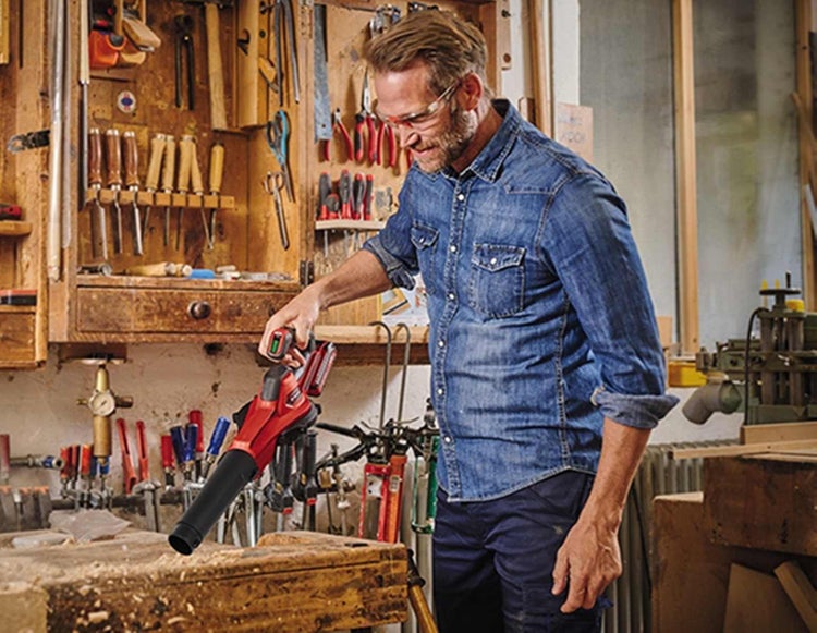 Ein Mann reinigt mit einem akkubetriebenen Handgebläse Holzspäne von einer Werkbank in einer gut ausgestatteten Werkstatt.