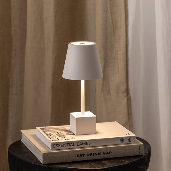 Moderne LED-Tischleuchte mit weißem konischem Schirm und quadratischem Fuß auf Büchern vor einem hellen Vorhang.