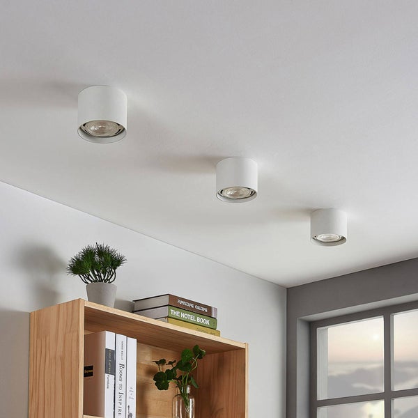 Drei zylindrische Aufbauspots an einer weißen Decke über einem organisierten Holzregal mit Büchern und Pflanzen in einem hellen, modernen Raum.