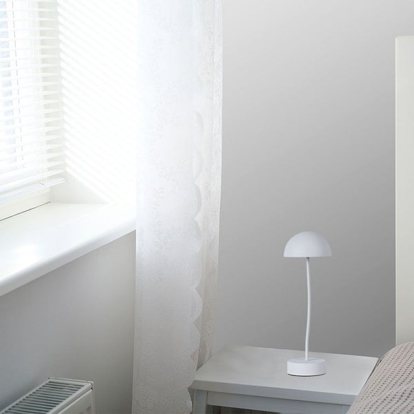 Weiße Tischleuchte auf einem Nachttisch neben einem Fenster mit Jalousien.