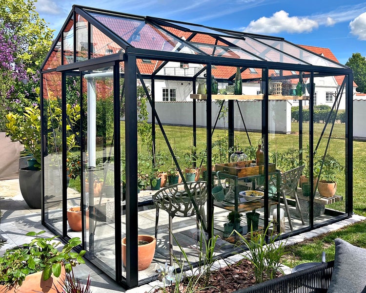 Modernes Gewächshaus mit schwarzem Aluminiumrahmen und Glaswänden, gefüllt mit Topfpflanzen und Gartenmöbeln in einem sonnigen Garten.