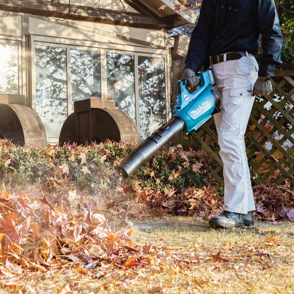 Eine Person benutzt einen Akku-Laubbläser von Makita, um Blätter in einem Garten zu entfernen.