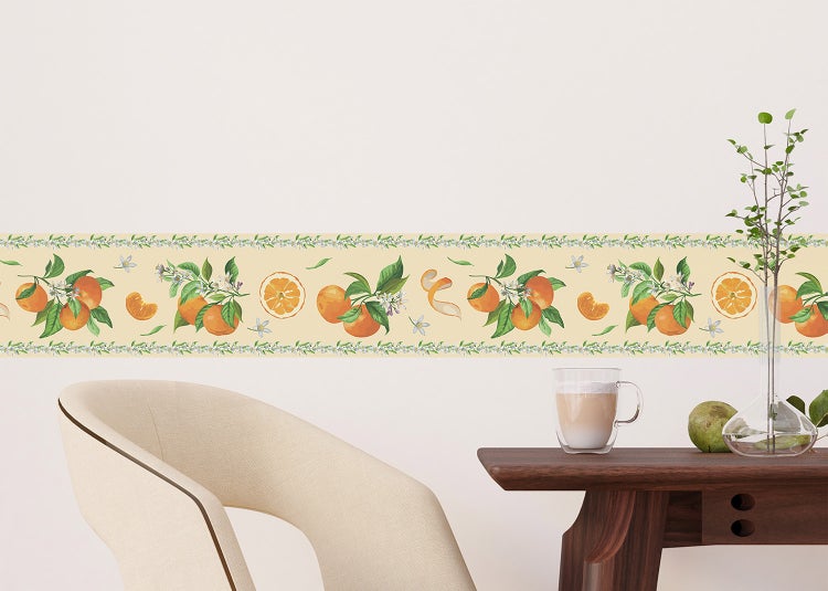 Tapetenbordüre mit Orangen-Motiv an einer hellen Wand über einem Holztisch und einem Sessel in einem hellen, mediterran gestalteten Raum.