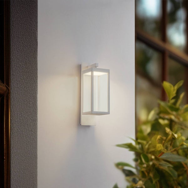 Moderne weiße Außenwandleuchte in Laternenform an einer Wand neben einem Fenster und Pflanzen.