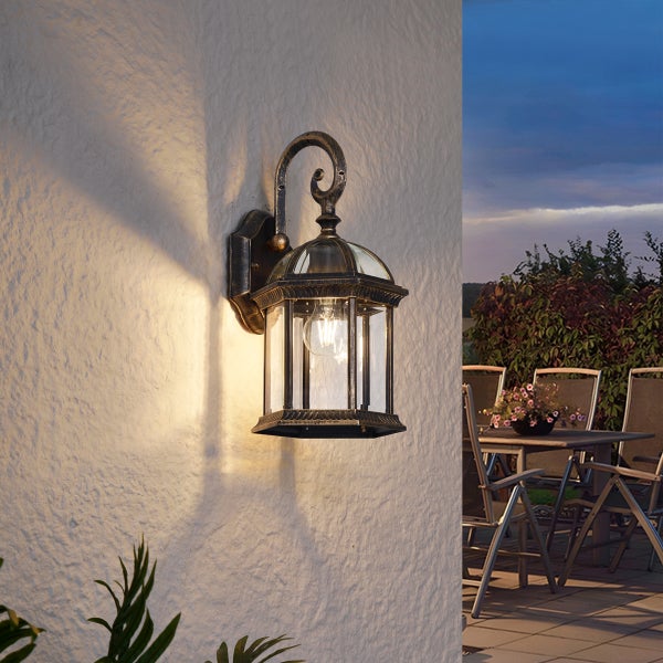 Klassische Außenwandleuchte im Laternendesign mit antikem Finish, montiert an einer strukturierten Wand neben einer Terrasse bei Dämmerung.