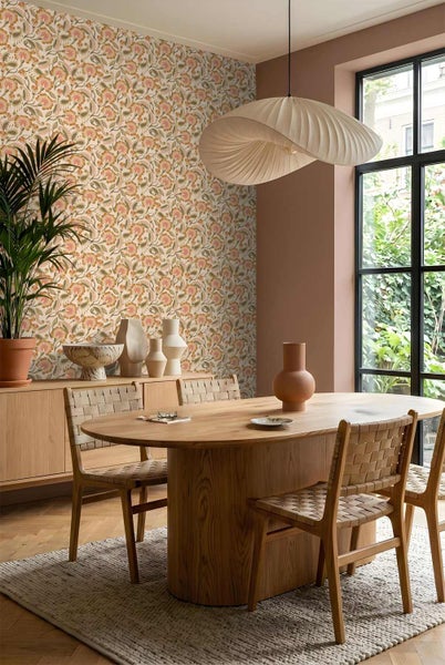 Esszimmer mit floral gemusterter Tapete, ovalem Holztisch und geflochtenen Stühlen unter einer großen Pendelleuchte.