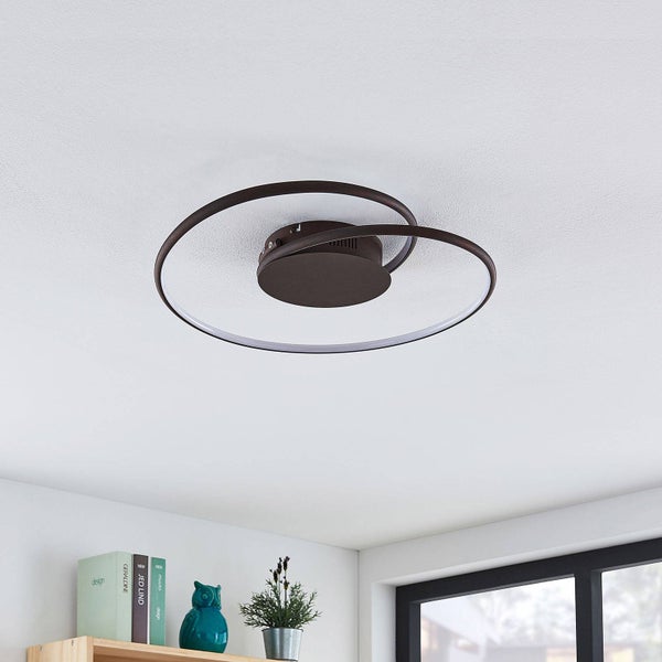 Moderne, runde LED-Deckenleuchte mit zwei überlappenden Ringen an einer Decke in einem hellen Raum mit Regal und Fenster.