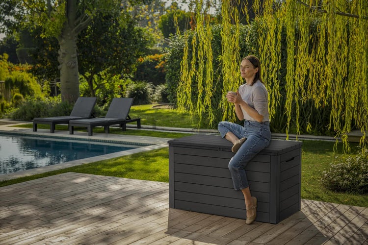 Große anthrazitfarbene Gartenbox in Holzoptik auf einer hellen Holzterrasse am Pool, genutzt als Sitzbank in einem hellen, gepflegten Garten.