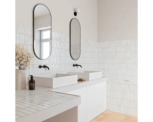 Modernes Badezimmer mit zwei Aufsatzwaschbecken, ovalen Spiegeln, schwarzen Wandarmaturen und vertikal verlegten hellen Fliesen.