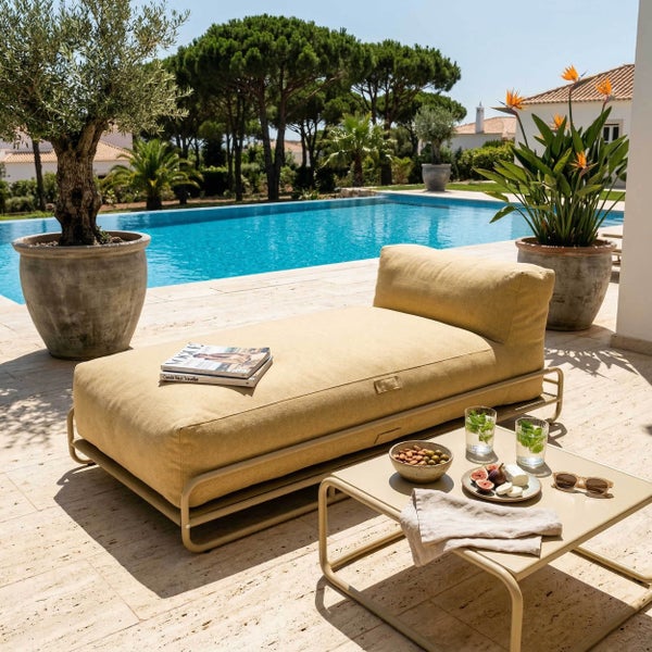 Beige Gartenliege und Beistelltisch aus Metall auf einer Steinterrasse neben einem Swimmingpool mit Pflanzen und Bäumen.