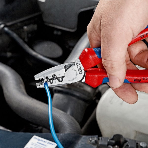 Eine Hand hält eine Kombinationszange, die ein blaues Kabel schneidet, vor einem Schaltschrank.