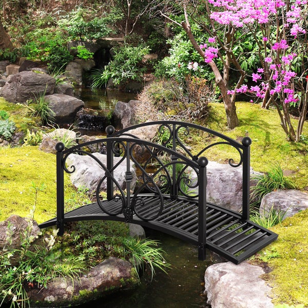 Dekorative Gartenbrücke aus Metall mit verschnörkeltem Geländer über einem kleinen Bachlauf in einer Gartenlandschaft mit Felsen und Pflanzen.