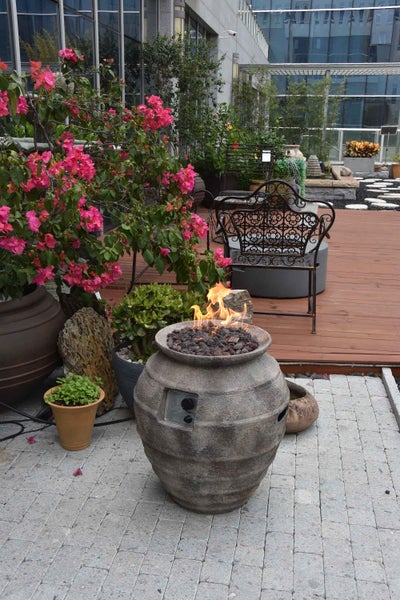 Gasfeuerstelle in Form einer rustikalen Steinvase mit brennender Flamme auf Lavasteinen auf einer gepflasterten Terrasse mit Pflanzen.