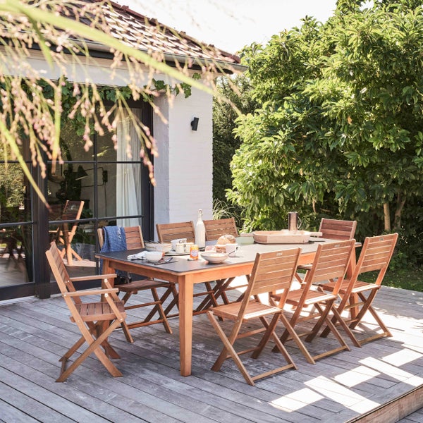 Moderne Gartenmöbel-Garnitur mit Holztisch und Klappstühlen auf einer Holzterrasse neben einem Haus, umgeben von Pflanzen.