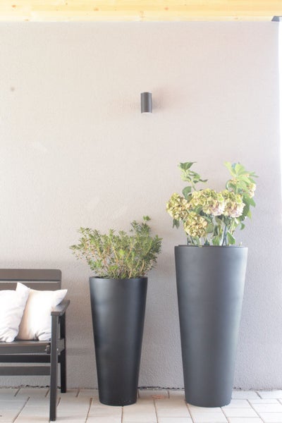Zwei hohe, konische Pflanzkübel in Anthrazit auf einer Terrasse vor einer grauen Wand, neben einer Gartenbank mit Kissen.