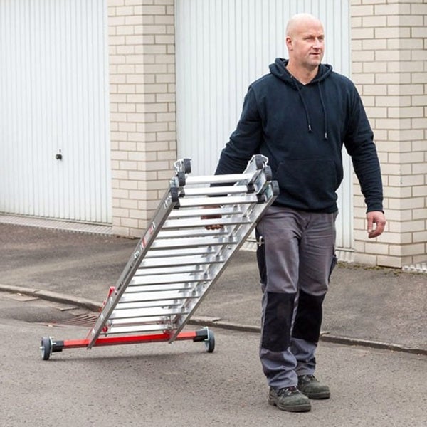 Ein Mann zieht eine zusammengeklappte Aluminium-Mehrzweckleiter mit montierten Rollen über einen befestigten Weg vor Garagentoren.