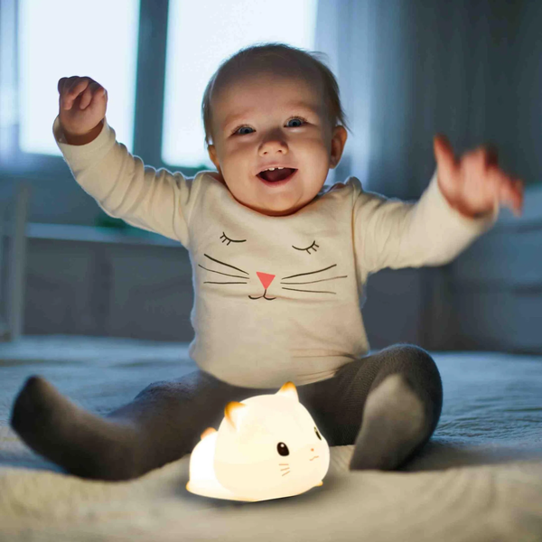 Ein lächelndes Kleinkind sitzt auf einem Bett neben einer leuchtenden, katzenförmigen LED Nachtlampe im Kinderzimmer.