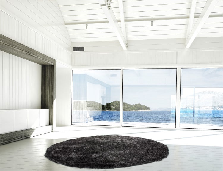 Heller Wohnraum mit holzverkleideten Wänden, Holzboden und einem runden Hochflor Teppich vor einer Fensterfront mit Blick auf das Meer.