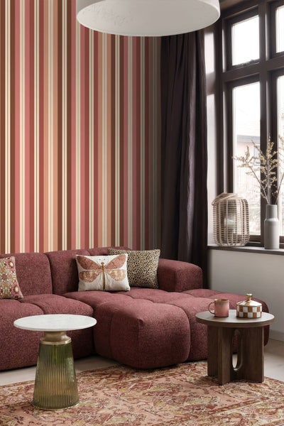 Wohnzimmer mit gestreifter Tapete in Rot- und Beigetönen, dunkelrotem Modulsofa mit Dekokissen und zwei kleinen Beistelltischen.