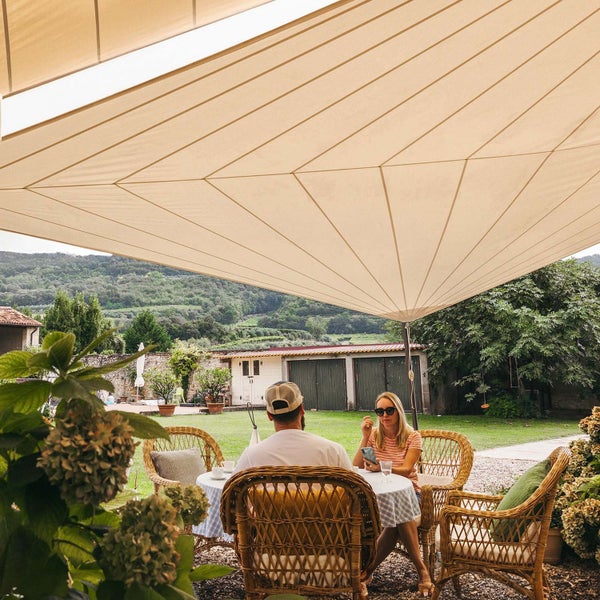 Großes beiges Sonnensegel über einer Terrasse mit Gartenmöbeln aus Korbgeflecht, einem Tisch und zwei Personen in einem weitläufigen Garten.