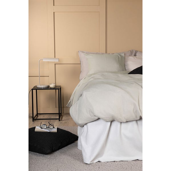 Schlafzimmer mit hellgrüner Bettwäsche, schwarzem Beistelltisch, weißer Tischlampe, Bodenkissen und Wandpaneelen.